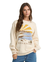 A woman modeling a cream-colored Billabong crewneck sweatshirt with a large graphic print of a sunset over waves and mountains, paired with blue jeans against a plain white background.