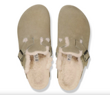 A pair of taupe suede clogs with a fluffy cream shearling lining, shown from a top-down view on a white background.