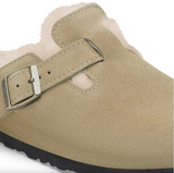 Close-up of a beige suede clog-style shoe with a soft light pink or cream shearling lining and a silver buckle strap, shown on a white background.