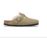 A single slip-on clog-style mule shoe in light tan suede with a soft cream shearling lining, a cork-look midsole, and a black outsole, shown in side profile on a white background.