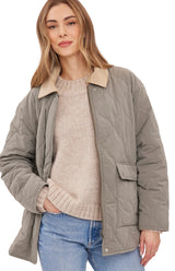 A woman wearing a quilted taupe-gray jacket with a beige collar, layered over a beige knit sweater and blue jeans, against a light gray studio background.