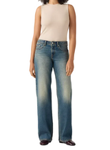 A model wearing a pair of medium-wash blue jeans with a faded light wash on the thighs and knees, paired with a beige sleeveless top and dark brown shoes, against a light gray studio background.
