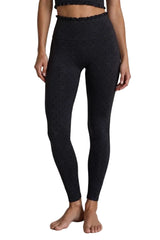 Black high-waisted leggings with a small repeating geometric pattern, modeled on a person standing barefoot against a light gray concrete wall and floor.
