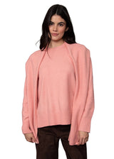 A woman modeling a light pink knit sweater with wide, draped sleeves and a relaxed fit, paired with dark brown pants, against a solid gray studio background.