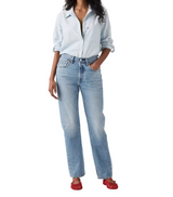 A model wearing light-wash blue denim jeans, a light blue button-down shirt, and red flat shoes, standing against a plain light gray studio background.