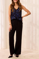 A woman modeling a sleeveless black top with a blue floral pattern and black wide-leg pants, standing in a brightly lit indoor setting with a light cream wall and wood floor.