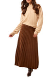 Woman wearing a beige sweater and brown pleated skirt against a white curtain background
