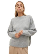 A woman wearing a light gray oversized knit sweater and beige trousers, standing against a plain light gray studio background.