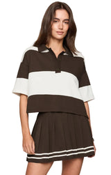 A woman modeling a color-blocked short-sleeve polo shirt and matching pleated mini skirt set. The set features dark brown and off-white horizontal stripes, with the skirt accented by double white stripes near the hem.