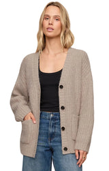 A woman wearing a beige textured knit cardigan with black buttons over a black tank top and blue jeans, photographed against a plain light beige studio background.